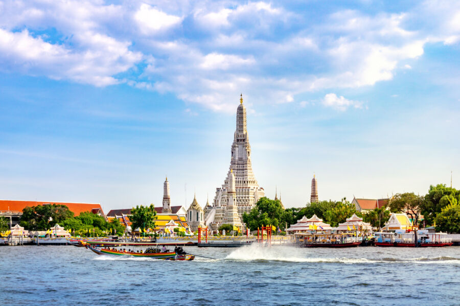 Scenic view of Wat Arun Temple in Bangkok, Thailand, featuring a long-tail boat navigating the river beside the iconic structure