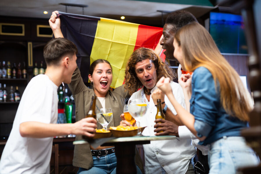 Diverse soccer fans celebrate Belgium's victory in a pub, holding a flag, pints of beer, and chips, filled with excitement