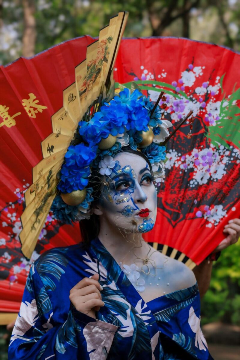 Colorful artistic portrait with theatrical makeup, ornate headdress, blue kimono, and vibrant red fan.