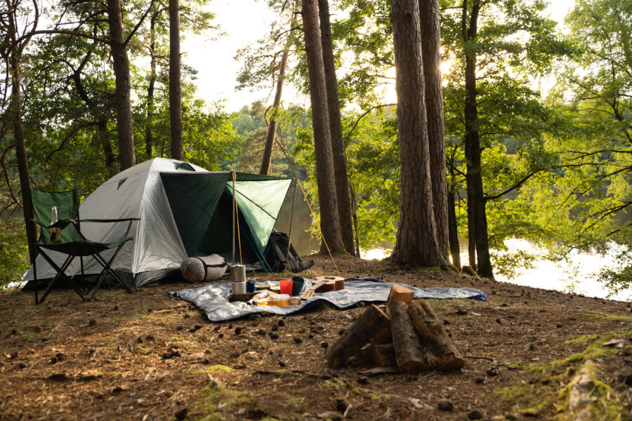 Tourist tent set up in a summer wood camping ground, featuring food and drinks arranged on a rug during the daytime.