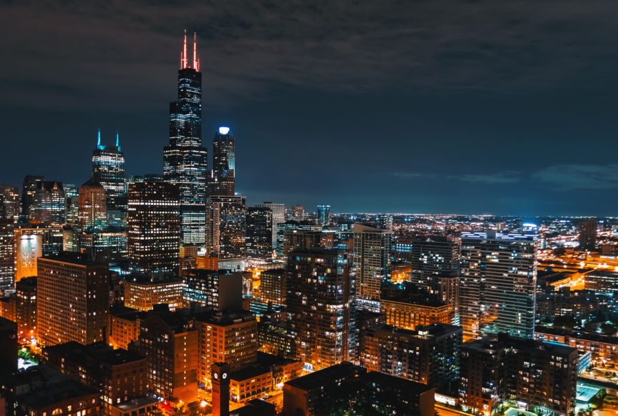 Nighttime cityscape of downtown Chicago, featuring tall skyscrapers glowing brightly against the evening backdrop