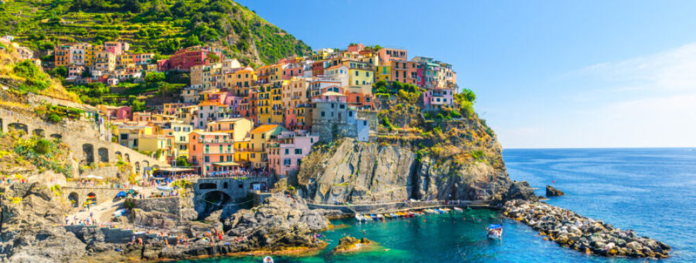 Manarola traditional typical Italian village in National park Cinque Terre, colorful multicolored buildings houses on rock cliff, fishing boats on water, blue sky background, La Spezia, Liguria, Italy