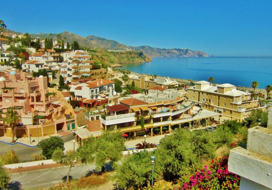 Picturesque Mediterranean seaside village with terracotta roofs, turquoise water, mountains, lush greenery.