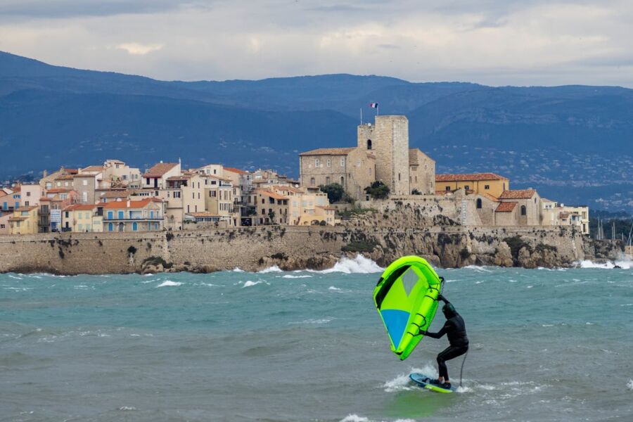 Windsurfer on vibrant waves near a Mediterranean coastal village with historic architecture.