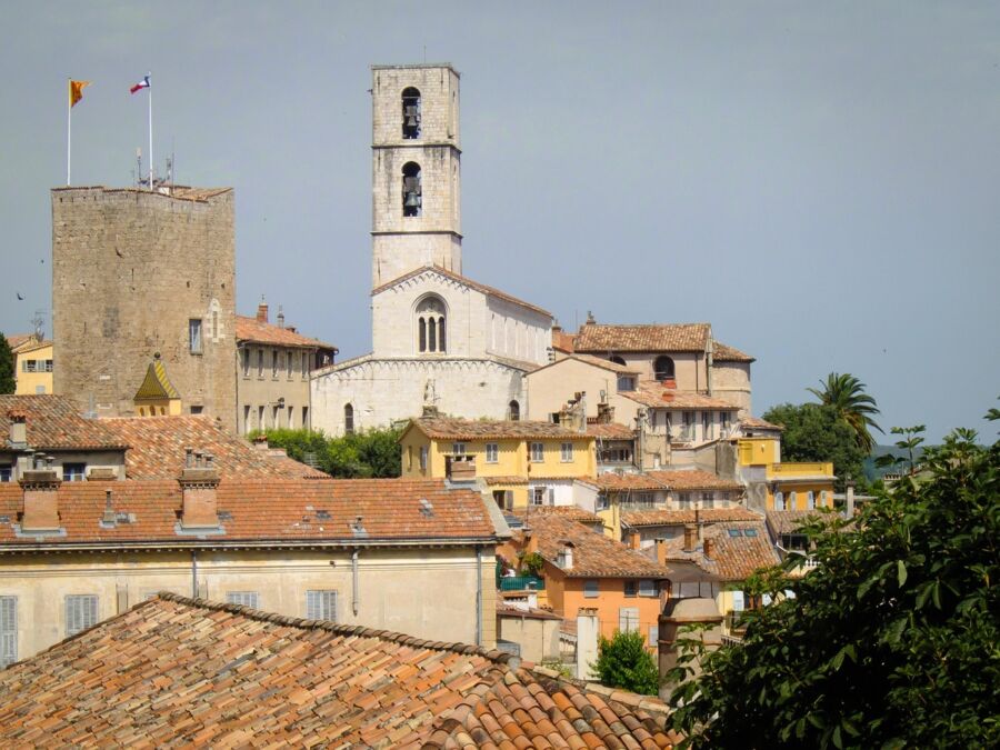 Historic European village with terracotta rooftops, stone tower, church, greenery, and waving flags.