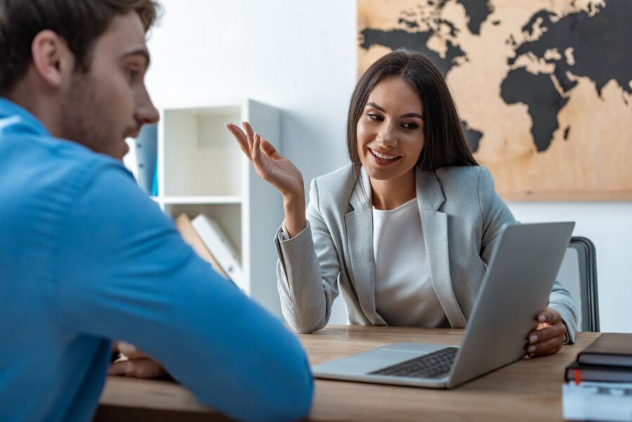 Friendly travel agent smiles as they discuss travel plans with a client, looking at a laptop screen