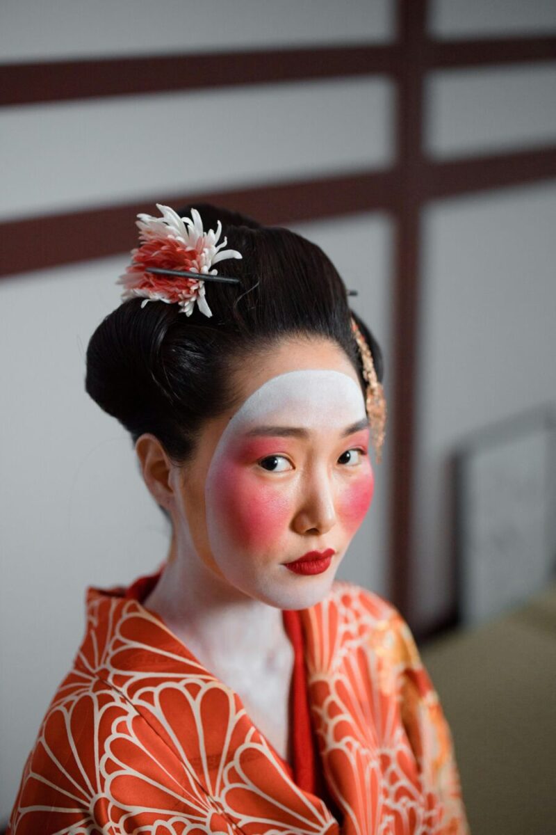 Traditional Japanese geisha with vibrant orange kimono, elaborate hairstyle, and floral kanzashi.