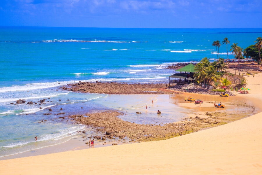 Panoramic view of Genipabu Beach in Brazil, showcasing golden sands and lush dunes under a clear blue sky