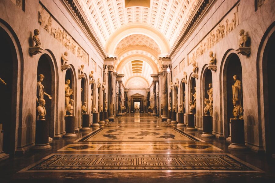 Grand museum hallway with classical architecture, vaulted ceiling, statues, marble floor, and mosaic designs.