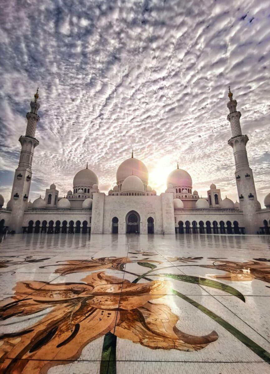 Grand mosque at sunset with white domes, minarets, floral mosaics, and reflective courtyard.