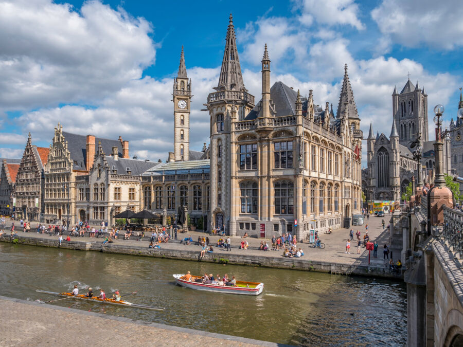 People enjoying a sunny day at Graslei quay by the river Lys in Ghent, with St Michael's Bridge in the background