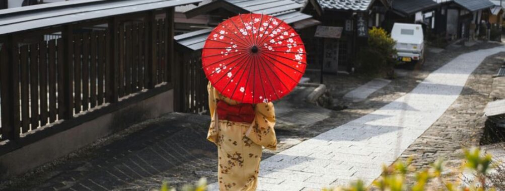 Traditional Japanese street scene with person in kimono, red parasol, and wooden buildings.