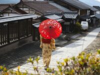 Traditional Japanese street scene with person in kimono, red parasol, and wooden buildings.