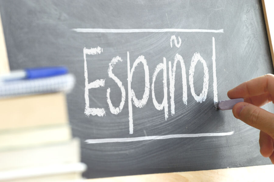 Handwriting on a blackboard in a language class, spelling "Spanish," with books and school materials nearby.