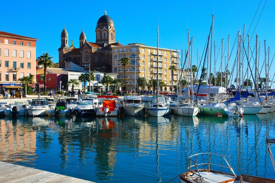 Vibrant marina with boats, neo-Romanesque church, palm trees, and sunny coastal promenade.