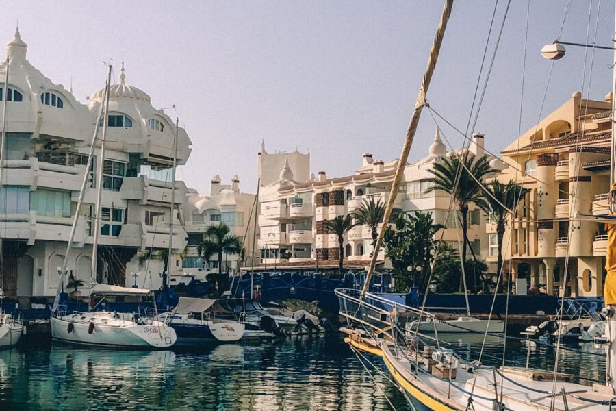 Scenic Mediterranean marina with sailboats, colorful architecture, palm trees, and serene waters.