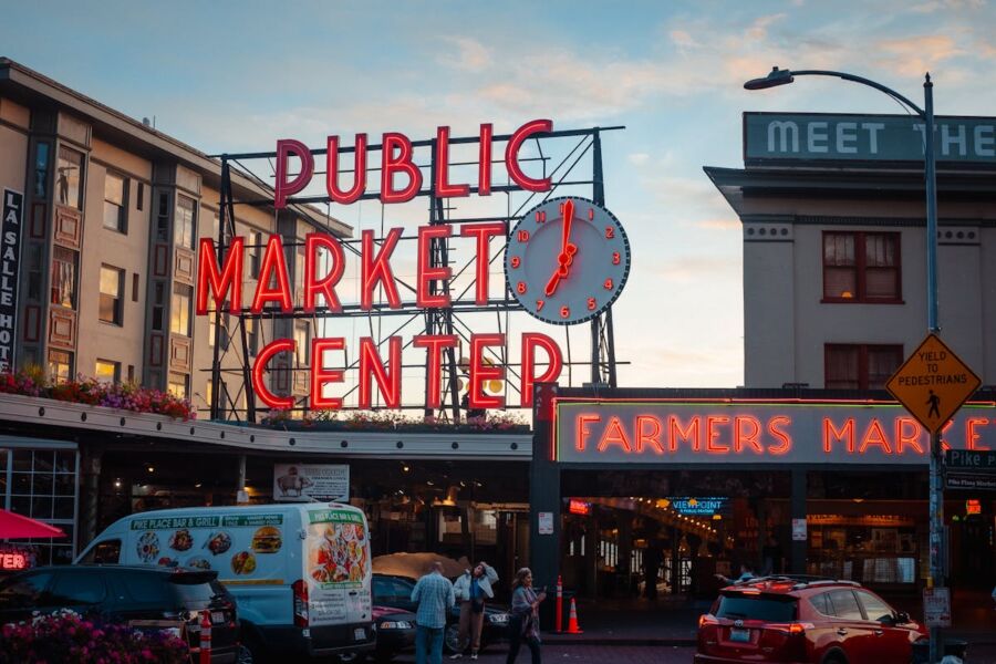 Pike Place Market neon sign at dusk, vibrant urban scene with pedestrians and food trucks.