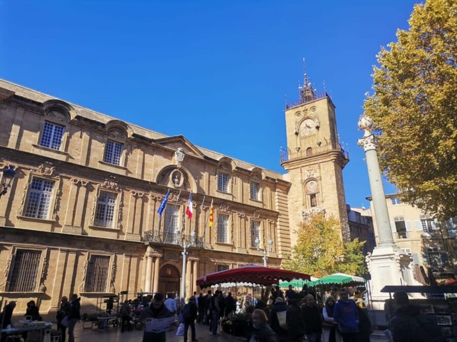 Historic town square with market stalls, ornate architecture, clock tower, and diverse visitors.