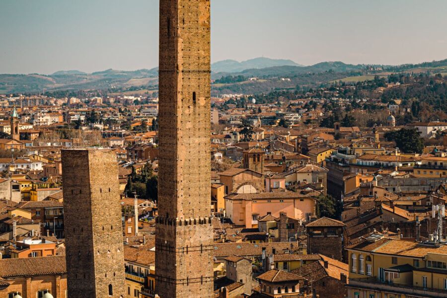 Medieval cityscape with historic towers, terracotta rooftops, lush hills, and clear blue sky.