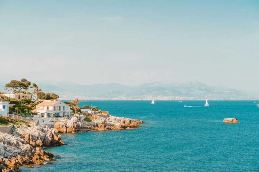 Serene coastal scene with turquoise waters, rocky shoreline, sailing boats, and hillside houses.