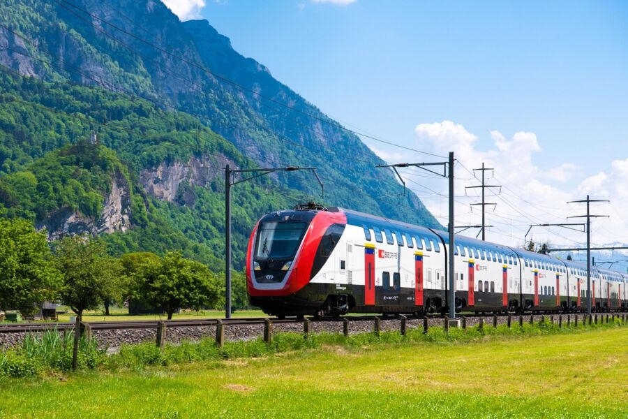 Modern red and white train traversing picturesque green landscape with mountains.