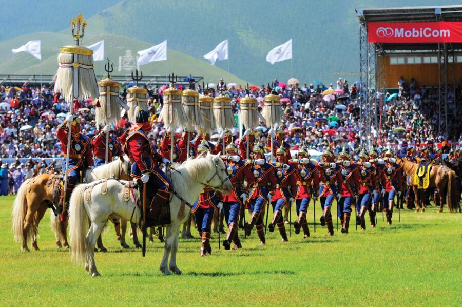 Mongolian festival riders in colorful attire on horses, with spectators and scenic hills.