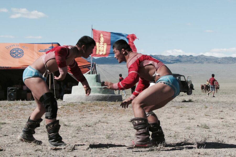 Mongolian wrestling match outdoors with traditional attire, cultural festival, mountains, and spectators.