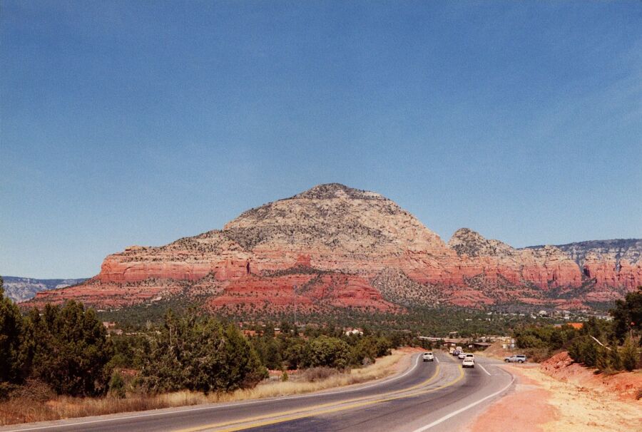 Majestic Sedona red rock formation with winding road, blue sky, and sparse greenery.