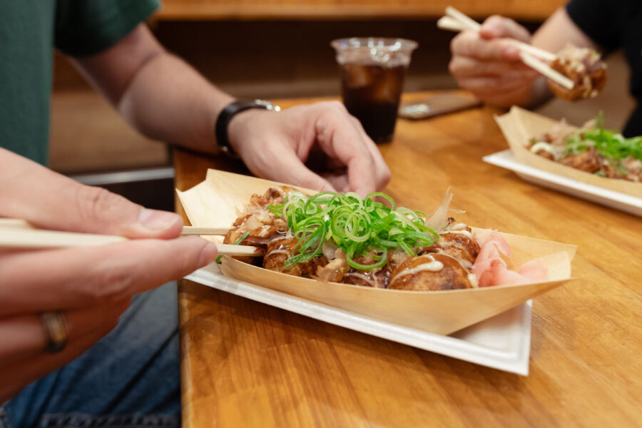 Man uses chopsticks to pick up a takoyaki ball in a bar, surrounded by friends, enjoying popular Osaka street food