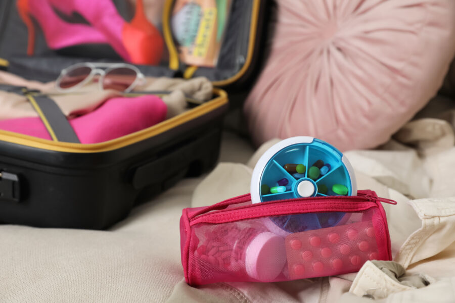 Medicines pouch filled with various medications and a pill box placed beside a packed suitcase on a sofa