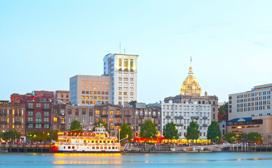 Panoramic view of Savannah, Georgia at sunset, showcasing the historic downtown skyline with illuminated buildings and steamboats.