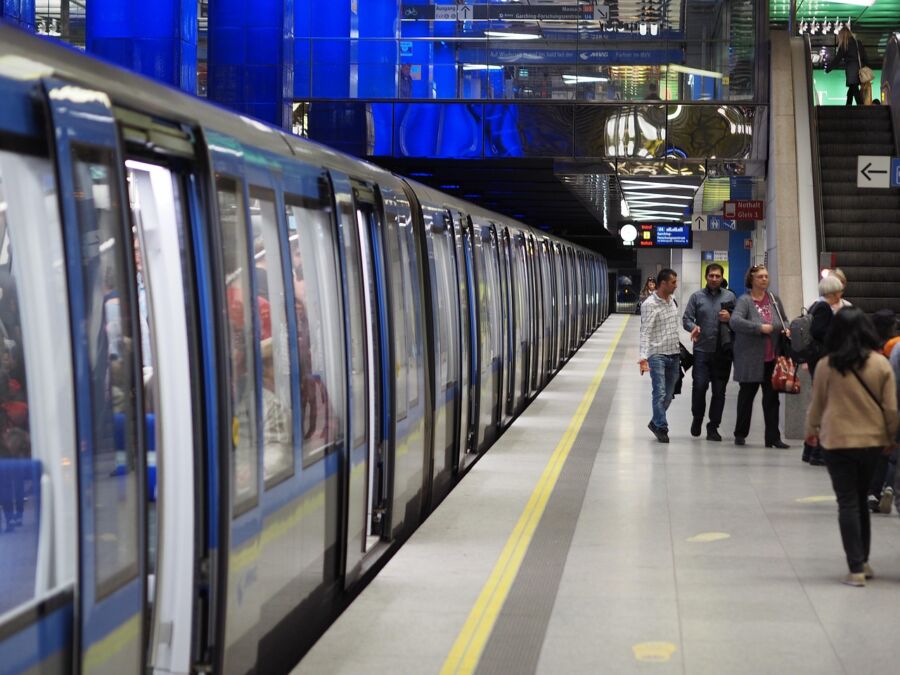 Munich sleek subway station features a blue-silver train with passengers visible through large windows. Commuters bustle on the well-lit platform, adorned with illuminated columns and digital displays, highlighting the modernity and efficiency of urban transit.