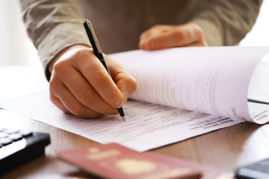 Close-up of a hand filling out forms with a pen, accompanied by a passport on a desk, showcasing travel documents