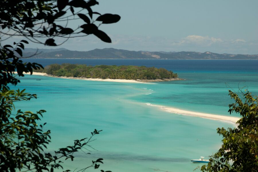 Tropical island view with lush foliage, sandy beach, and clear blue waters under a sunny sky.