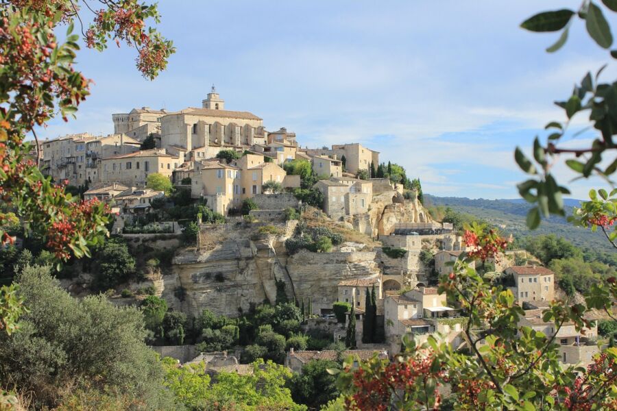 Village on rocky cliff with earthy buildings and church, surrounded by lush greenery and rolling hills.