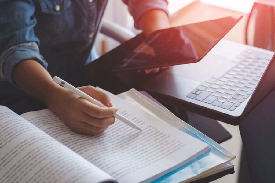 Young woman studying at home, using a digital tablet and laptop while reading a book